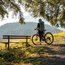 Cyclist with helmet and backpack stands in forest beside bench with mountain view